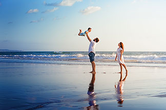Schwimmzubehör in Top-Qualität: Eine junge Familie steht am Strand. Der Vater hebt das Kind in die Luft hoch.