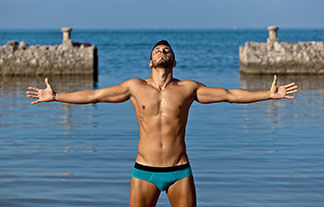 Ein junger, sportlicher Mann in einem Slip genießt mit ausgestreckten Armen die Sonne am Strand.