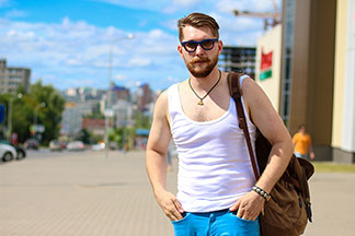 Ein junger Mann, der ein weißes Herrenunterhemd, eine Sonnenbrille und einen Rucksack trägt, schlendert lässig durch eine Stadt.