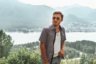 Ein junger Mann mit einem karierten, offen getragenen Kurzarmhemd steht vor einem See mit Bergen im Hintergrund.