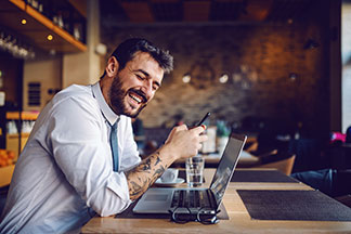 Ein bärtiger Mann mit Tattoos sitzt in einem hellblauen Herrenhemd in einem Café vor seinem Laptop und lacht.