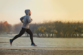Ein junger Mann joggt am frühen Morgen in einer grauen Fleecejacke mit Kapuze durch einen herbstlichen Park.