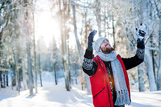 Ein bärtiger Mann, der eine Mütze, Handschuhe und einen Winterschal trägt, steht in einem verschneiten Wald und streckt die Arme Richtung Himmel.