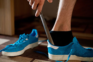 Sneakersocken für Herren: Nahaufnahme der Beines eines Mannes, der schwarze Sneakersocken trägt, und sich gerade mit einem Schuhlöffel einen blauen Turnschuh anzieht.