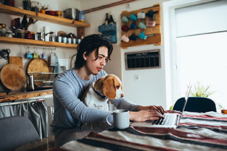 Ein junger Mann, der Homewear trägt, arbeitet an seinem Küchentisch an seinem Laptop und hat seinen Hund auf dem Schoß.