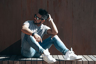 Herrenschuhe für die Freizeit: Ein junger Mann, der weiße Turnschuhe, Jeans, ein graues Shirt und eine Sonnenbrille trägt, sitzt vor einer braunen Wand.