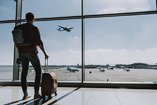 Ein Mann steht am Flughafen mit seinem Koffer-Trolley und schaut aus dem Fenster.