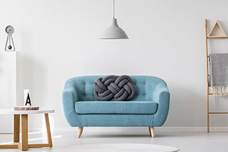 Mittig im Raum steht eine türkisfarbene Couch, über der eine eine graue Deckenlampe hängt – links im Zimmer steht ein runder Couchtisch mit weißer Tischplatte, an der Rückwand leiterförmiges Regal.