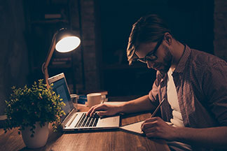 Tischleuchte am Schreibtisch: Ein junger Mann sitzt mit einem Stift und einem Block vor einem Laptop an einem gut ausgeleuchteten Holzschreibtisch – um ihn herum ist es dunkel.