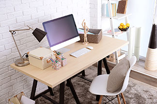 Ordnung mit der richtigen Büroausstattung: Ein Schreibtisch mit einer hellen Tischplatte aus Holz auf dem eine Schreibtischlampe, ein Monitor und verschiedene Ordnungskörbe stehen.