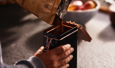 Eine silberne Aromadose von Tchibo wird von einer linken Hand gehalten, während eine rechte Hand gemahlenen Kaffee mit einem Kaffeelot entnimmt.