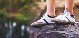 Sportliche Herrenstrümpfe: Nahaufnahme der Beine eines jungen Mannes, der weiße Sneakers und schwarze Sneakersocken trägt, während er auf einem Felsen steht.