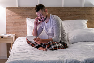 Ein junger Mann sitzt abends in seinem Schlafanzug – mit Bündchen an den Beinen und Knopfleiste am Oberteil – auf seinem Bett und telefoniert, während er in sein Tablet schaut..