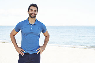 Sportshirt für den Alltag: Ein bärtiger Mann in einem hellblauen Poloshirt lächelt fröhlich an einem Strand in die Kamera.