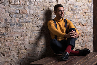 Ein junger Mann mit Brille sitzt an eine Steinwand gelehnt im Schneidersitz und trägt rote Herrensocken mit schwarzen Schuhen.
