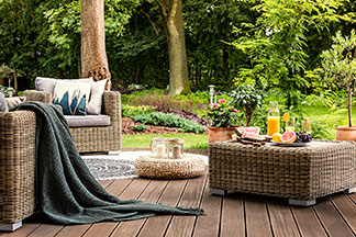 Urlaub im Garten: Eine Holzterrasse mit Gartenmöbeln aus Rattan in einem grünen Garten mit hohen Bäumen.