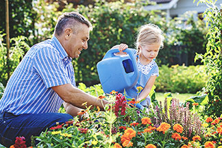 Blütenpracht: Ein Vater kniet vor einem Blumenbeet im Garten, seine kleine Tochter steht dabei und gießt mit einer blauen Gießkanne die Blumen.