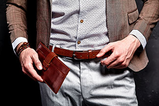 Nahaufnahme eines Mannes, der einen braunen Herrengürtel aus Leder trägt, sowie in der Hand den passenden braunen Geldbeutel hält.