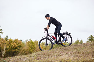 Ein junger Mountainbiker mit Mütze und langer, schwarzer, enganliegender Fahrrad-Sporthose fährt durch eine hügelige Landschaft.