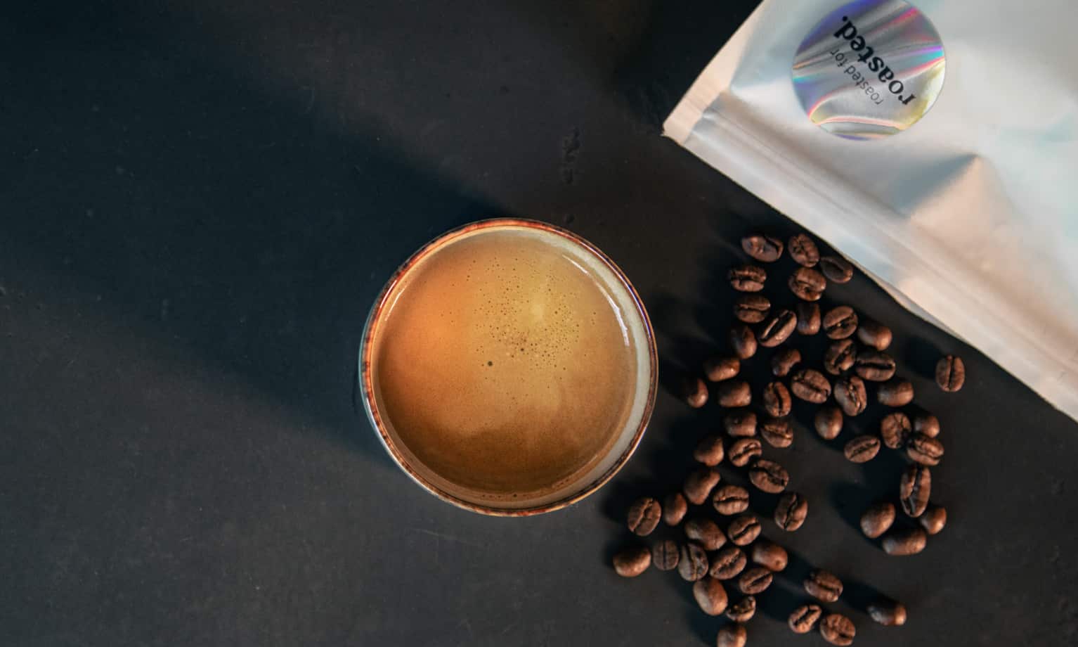Auf einem Tisch steht eine Tasse Co-Roast #1 Espresso auf dem Kaffeebohnen überall verteilt sind