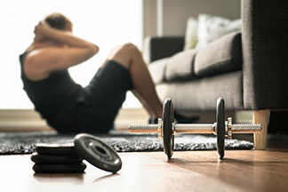 Training daheim: Ein junger Mann macht Sit-ups in seinem Wohnzimmer. Vor ihm liegen Hanteln als Fitness-Zubehör.