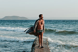 Ein junger Mann, der eine Badehose, eine Sonnenbrille und einen offenen, beigen Bademantel trägt, läuft auf einem steinernen Steg, der in Meer reicht.