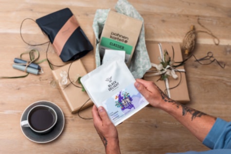 Jemand hält eine Packung Kaffee in den Händen während auf dem Tisch darunter zwei weitere Packungen und Geschenkpapier liegen