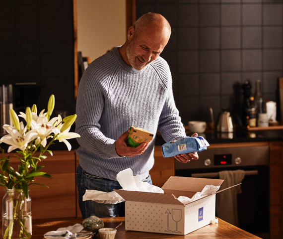 Älterer Mann packt in einer Küche ein Tchibo Paket mit Kaffee aus und hält in der einen eine Packung Feine Milde und in der anderen Hand den Bio Kaffee.
