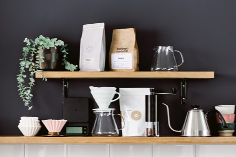 Zwei Regalbretter sind an einer dunkelgrauen Wand befestigt und es stehen drei Packungen Specialty Coffee darauf sowie verschiedene Kaffeezubereiter und eine Pflanze.