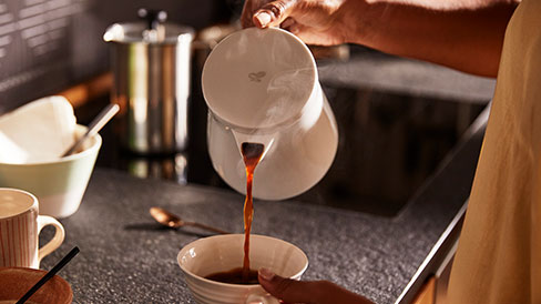 Frau gießt Kaffee aus einem Porzellan-Kaffeebereiter mit Permanentfilter in eine Tasse