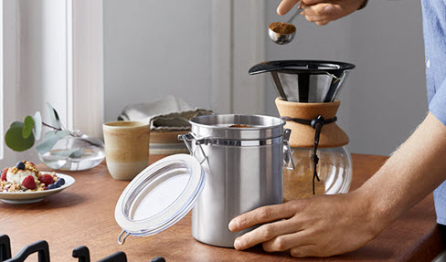 Eine silberne Aromadose von Tchibo wird von einer linken Hand gehalten, während eine rechte Hand gemahlenen Kaffee mit einem Kaffeelot entnimmt.