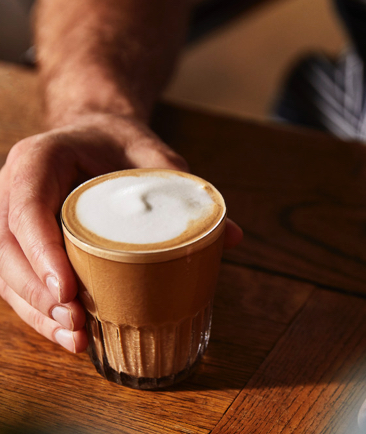 Ein Glas Milchkaffee wird von einer Hand auf einem Holztisch festgehalten