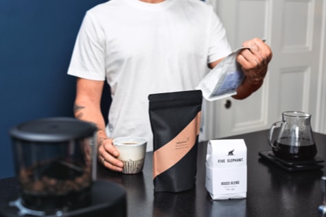 Jemand hält eine Packung Specialty Coffee in der einen Hand und eine Tasse Kaffee in der anderen während auf dem Tisch darunter zwei weitere Packungen und eine Moccamaster steht