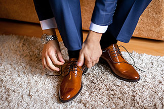 Herrenschuhe fürs Büro: Nahaufnahme der Füße eines Mannes, der einen blauen Anzug trägt, auf seinem Bett sitzt und sich seine braunen Lederschuhe bindet.