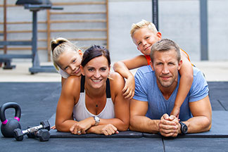 Sportaccessoires für die ganze Familie: Ein Mann und eine Frau liegen mit dem Bauch auf grauen Sportmatten, auf ihren Rücken liegen der Sohn und die kleine Tochter.