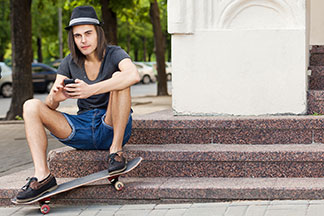 Ein stylisch gekleideter Skater, der einen dunklen Hut und eine Jeans-Shorts trägt, sitzt lässig auf einer Marmortreppe und bedient sein Smartphone.