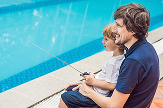 Ein Vater, der ein blaues Polohemd trägt, sitzt mit seinem Sohn am Rand eines Swimmingpools. Beide lenken gemeinsam ein ferngesteuertes Boot.