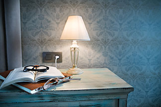 Schickes Design für Ihre Lampe: Eine Tischleuchte mit einem weißen Lampenschirm steht auf einem Tisch in Vintage-Optik, auf dem eine Mappe, ein aufgeschlagenes Buch und eine Brille liegen.