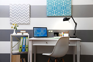 Arbeitsplatz richtig dekorieren: Ein weißer Schreibtisch mit Lampe und Monitor steht vor einer horizontal grau-weiß gestreiften Wand an der zwei bunte Dekobilder hängen.