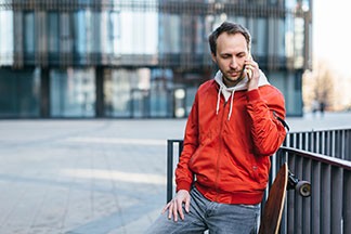 Sportjacke für den Alltag: Ein junger Mann, der eine rote sportliche Jacke trägt und ein Longboard bei sich hat, telefoniert mit seinem Smartphone.