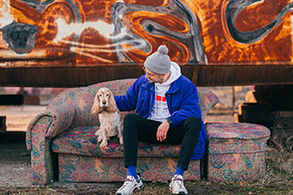 Ein junger Mann, der passend zu seiner blauen Jacke blaue Herrenstrümpfe trägt, sitzt im Freien mit seinem Hund auf einer alten Couch.