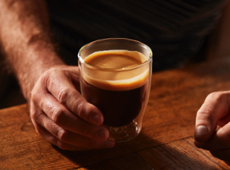 Mann hält ein auf dem Tisch stehendes Caffè Crema Glas mit Kaffee fest.