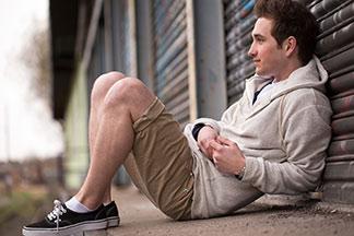 Ein Mann mit Sweatshirt-Jacke und beiger Bermuda sitzt an draußen an eine Wand gelehnt