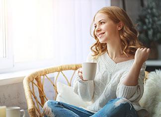 Eine Frau im weißen Pullover sitzt in ihrem Wohnzimmer und genießt eine Tasse Kaffee.