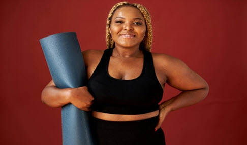 Passt perfekt: Eine Frau trägt einen schwarzen Sport-BH in einer großen Größe und hält eine Yoga-Matte unter dem Arm.