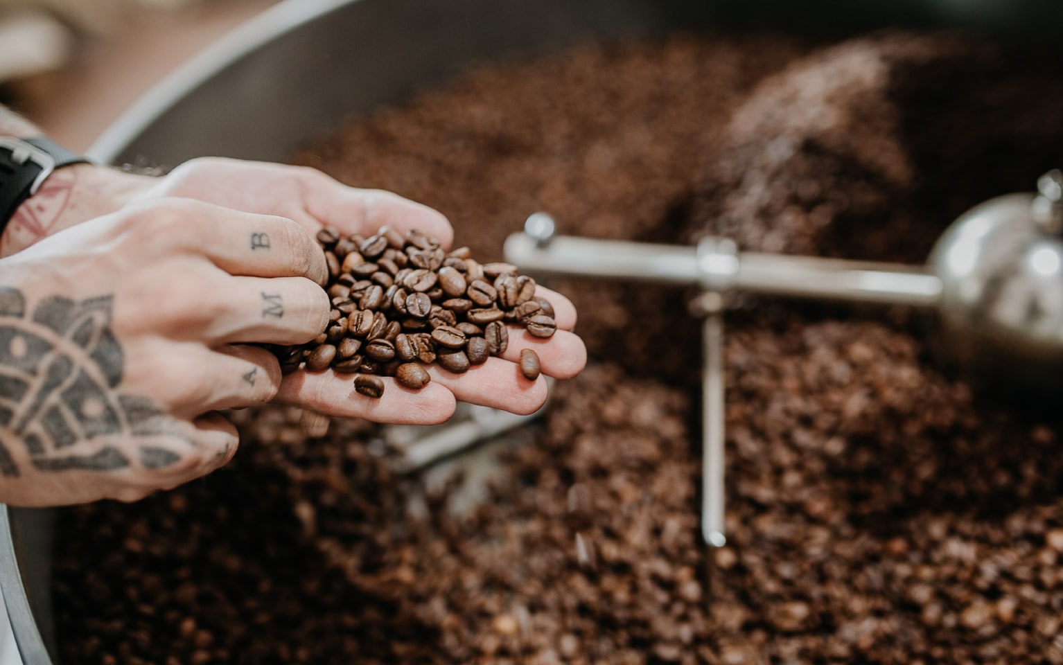 Eine tätowierte Hand nimmt Kaffeebohnen aus einer Röstertrommel