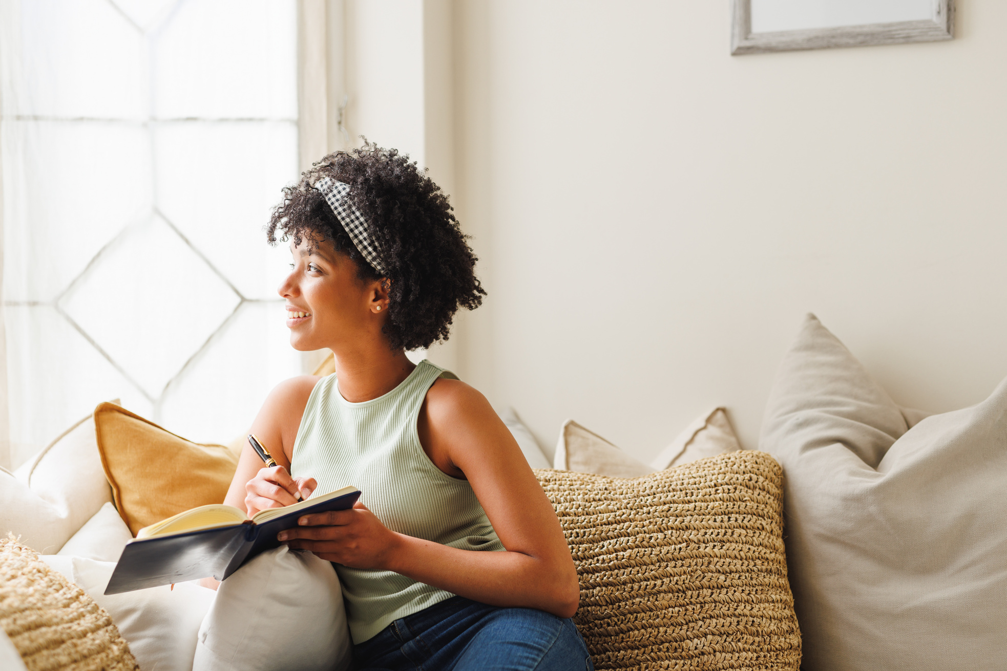 Nachdenkliche junge Frau mit Afrohaar, die aus dem Fenster schaut und auf dem Sofa in ihr Tagebuch schreibt.