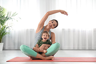 Yoga-Kleidung in Top-Qualität: Eine junge Mutter sitzt auf ihrer Yogamatte und hebt den Arm über den Kopf, Ihr Sohn sitzt auf ihrem Schoß.