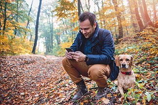 Ein Mann in einer blauen Übergangsjacke und mit wetterfesten Herren-Stiefeln kniet in der Hocke in einem herbstlichen Wald und bedient sein Smartphone, neben ihm liegt sein Hund.
