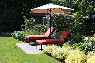 Zwei Gartenliegen mit einer roten Auflage stehen nebeneinander in einem Garten – dazwischen ein aufgespannter Sonnenschirm.